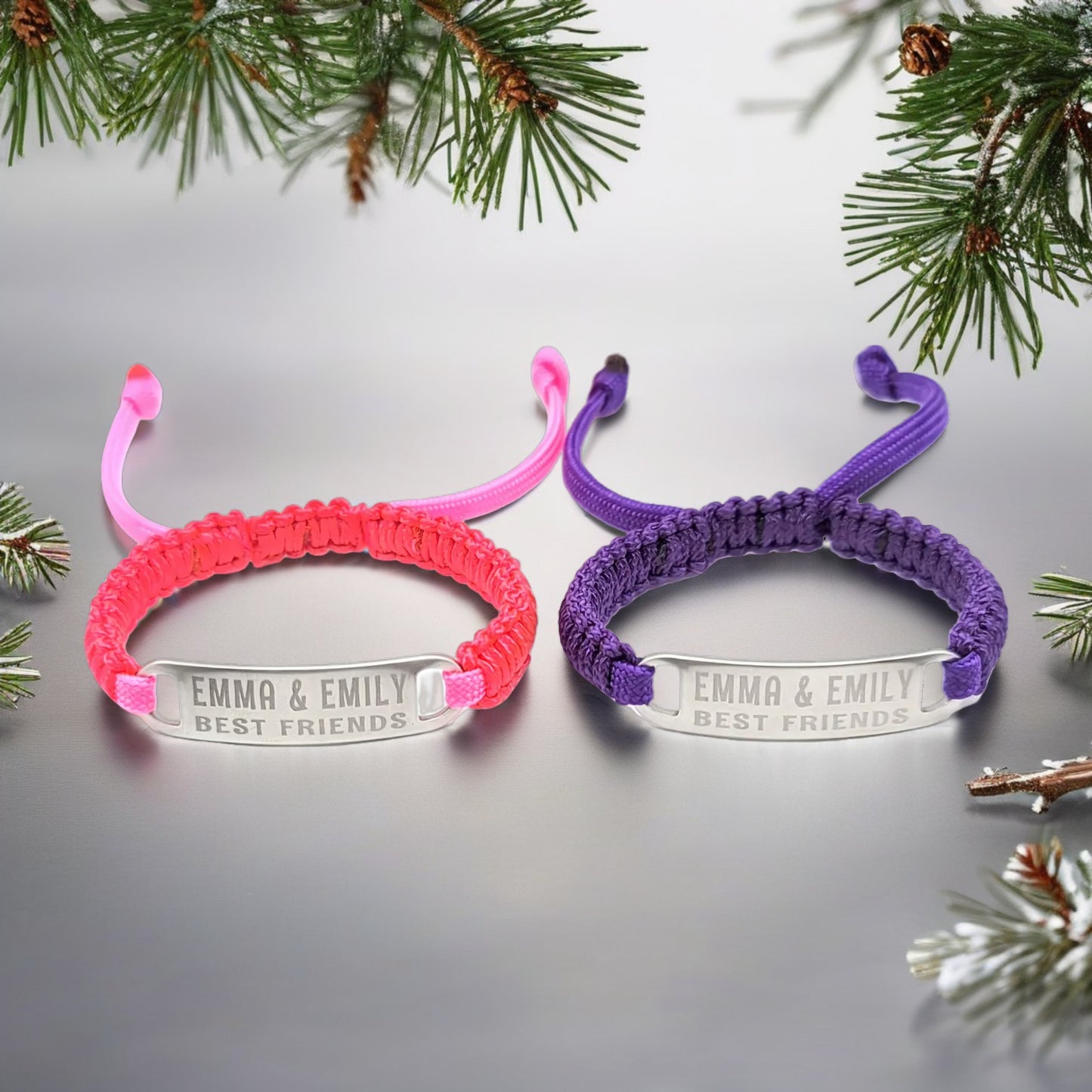 A close-up of the purple and pink adjustable micro paracord bracelets with engraved tags that read Emma & Emily Best Friends, displayed on a flat surface surrounded by holiday-themed pine branches.