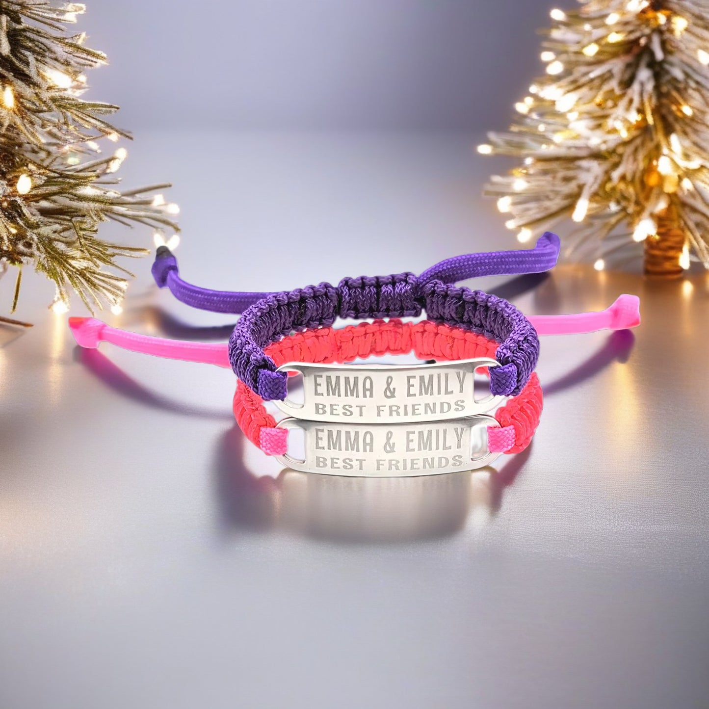 Purple and pink best friends adjustable paracord bracelets stacked together, displayed upright, with Emma & Emily Best Friends engraved on both tags. The background features a lit holiday tree and a festive atmosphere.