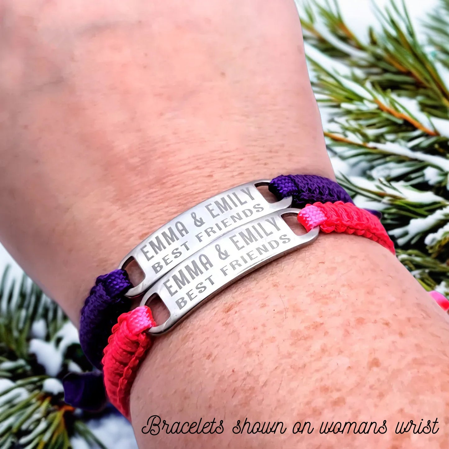 A woman's wrist wearing both the purple and pink adjustable best friends bracelets, engraved with Emma & Emily Best Friends, shown in a winter-themed setting with pine branches.