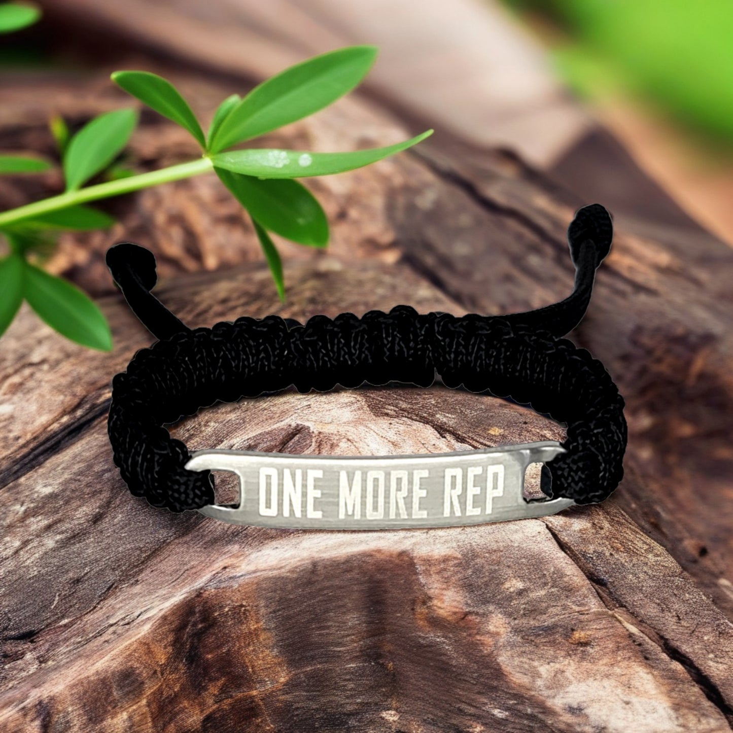 A close-up of a black micro cord bracelet with "One More Rep" engraved on a metal tag, lying on a wooden surface with green leaves nearby.