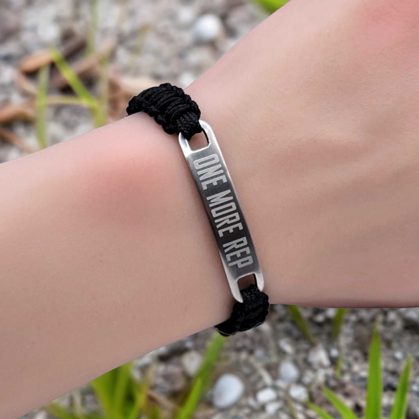 A black micro cord bracelet with the words "One More Rep" engraved on a metal tag, worn on a wrist with a natural background of grass and small stones