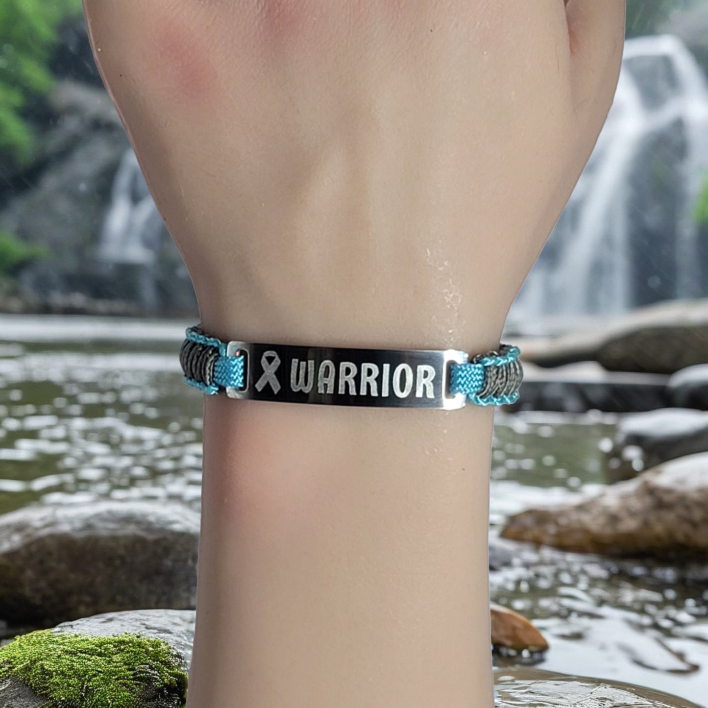 A bracelet with teal and black micro cord on a wrist featuring an engraved metal tag that says Warrior with a ribbon symbol displayed near a calm waterfall background.