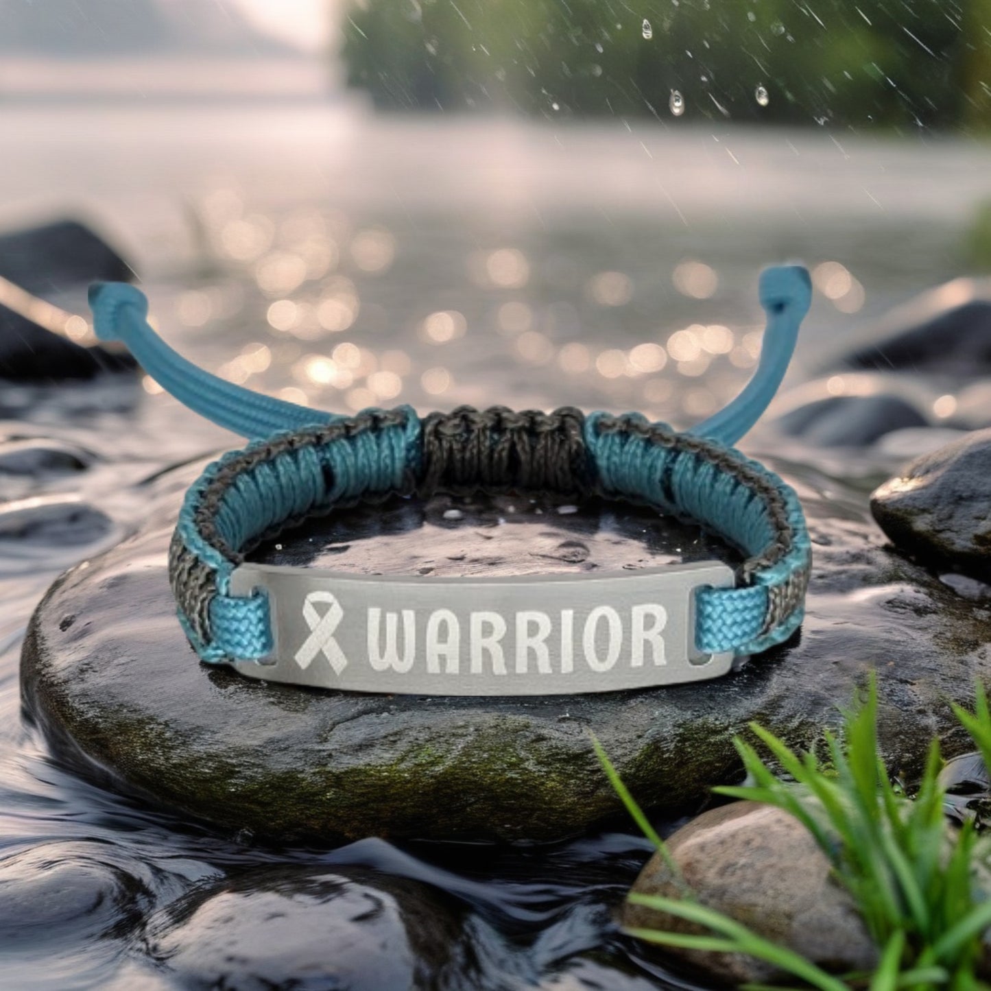 A close-up of a bracelet with teal and black micro cord featuring a metal tag engraved with the word Warrior and a ribbon symbol positioned on a wet rock by a peaceful lake with raindrops falling.