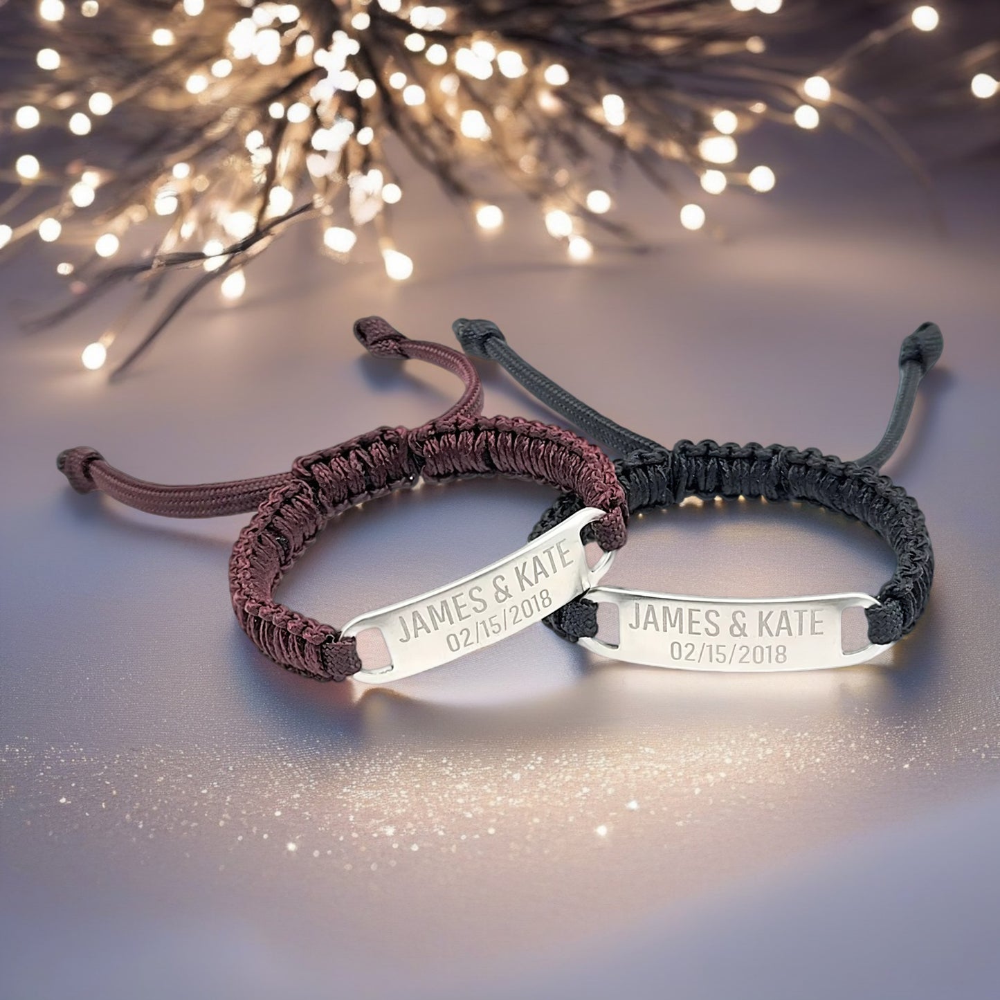 A romantic-themed display of two micro paracord bracelets, one in maroon and one in black, with engraved text. The bracelets are set against a soft, sparkling backdrop with a focus on the polished tags.