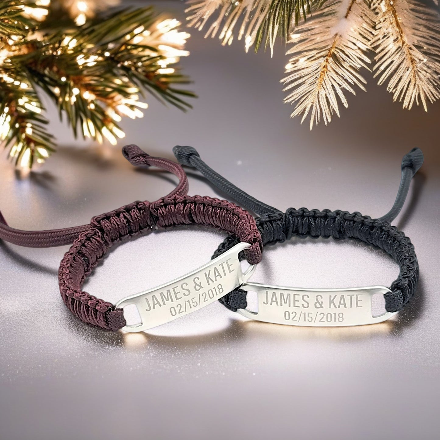 Two engraved micro paracord bracelets (maroon and black) displayed under a festive pine tree, with warm lighting and a soft-focus holiday ambiance. The engraved names and date are clearly visible.