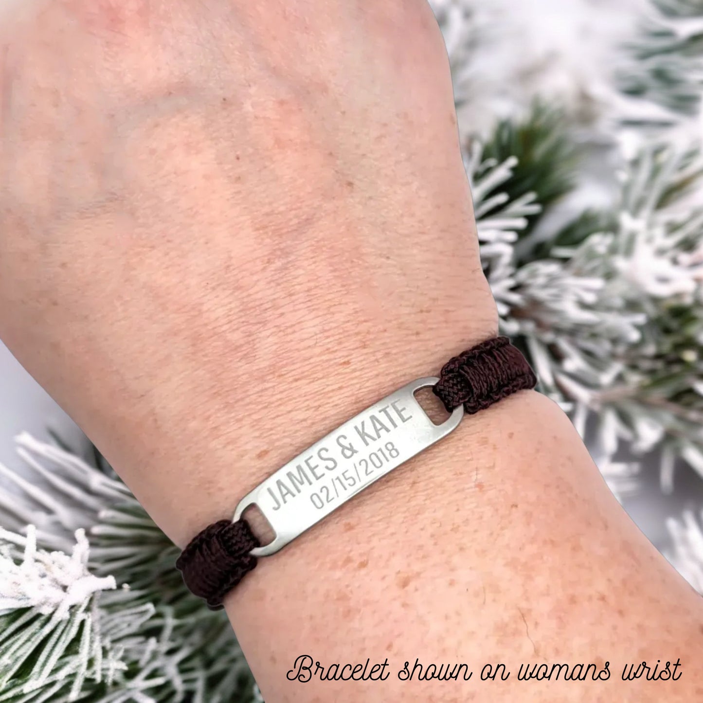 A maroon micro paracord bracelet shown on a woman's wrist, emphasizing the engraved stainless steel plate reading "James & Kate 02/15/2018." The background features frosted pine branches for a winter-themed presentation.