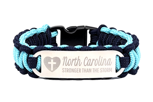 Navy blue and light blue thin 275 paracord ID bracelet with a silver, stainless steel ID tag engraved with North Carolina Stronger than the Storm and a cross in a heart symbol, displayed on a white background.