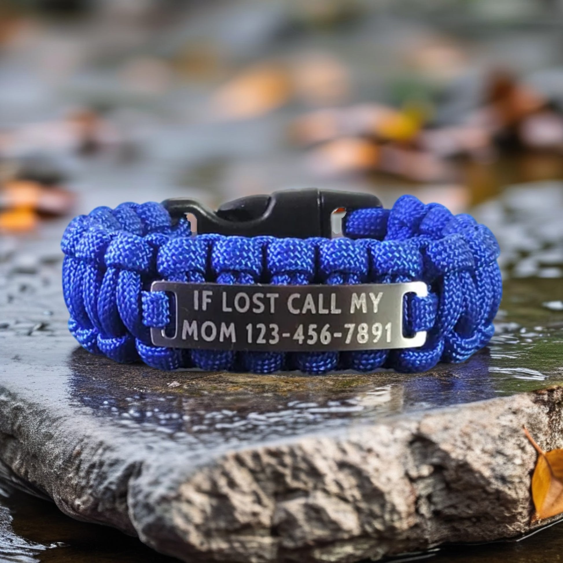  A bright blue paracord bracelet with a polished tag engraved with the text If Lost Call My Mom followed by a phone number resting on a wet stone with blurred foliage in the background.