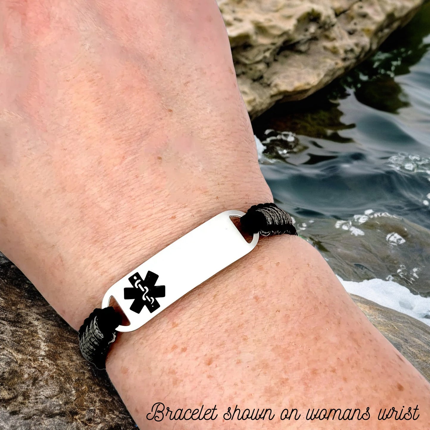 Adjustable micro paracord medical ID bracelet shown on a womans wrist near a rocky stream. Features a black and gray cord with a polished silver tag engraved with a black medical alert symbol, highlighting its fit and design.