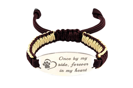 An adjustable micro paracord bracelet displayed on a clean white background. The bracelet features a polished stainless steel oval tag engraved with the phrase Once by my side, forever in my heart, accompanied by a paw print and heart design. The paracord is intricately woven in maroon and tan tones, creating a minimalistic and elegant look