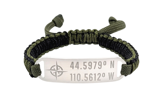 Adjustable micro paracord coordinate bracelet on a white background. Features black and olive green cords with a polished silver tag engraved with coordinates and a compass symbol, showcasing a personalized and functional design.