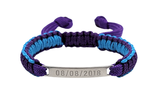 An adjustable micro paracord bracelet displayed on a white background. The bracelet features a polished mini tag engraved with a date and a paracord weave in purple and teal.