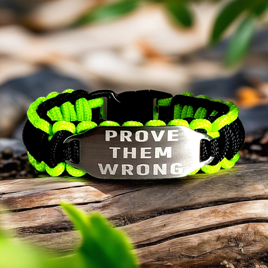 A neon green and black paracord bracelet with an engraved tag that says "Prove Them Wrong," displayed on a wooden surface with blurred natural background elements.