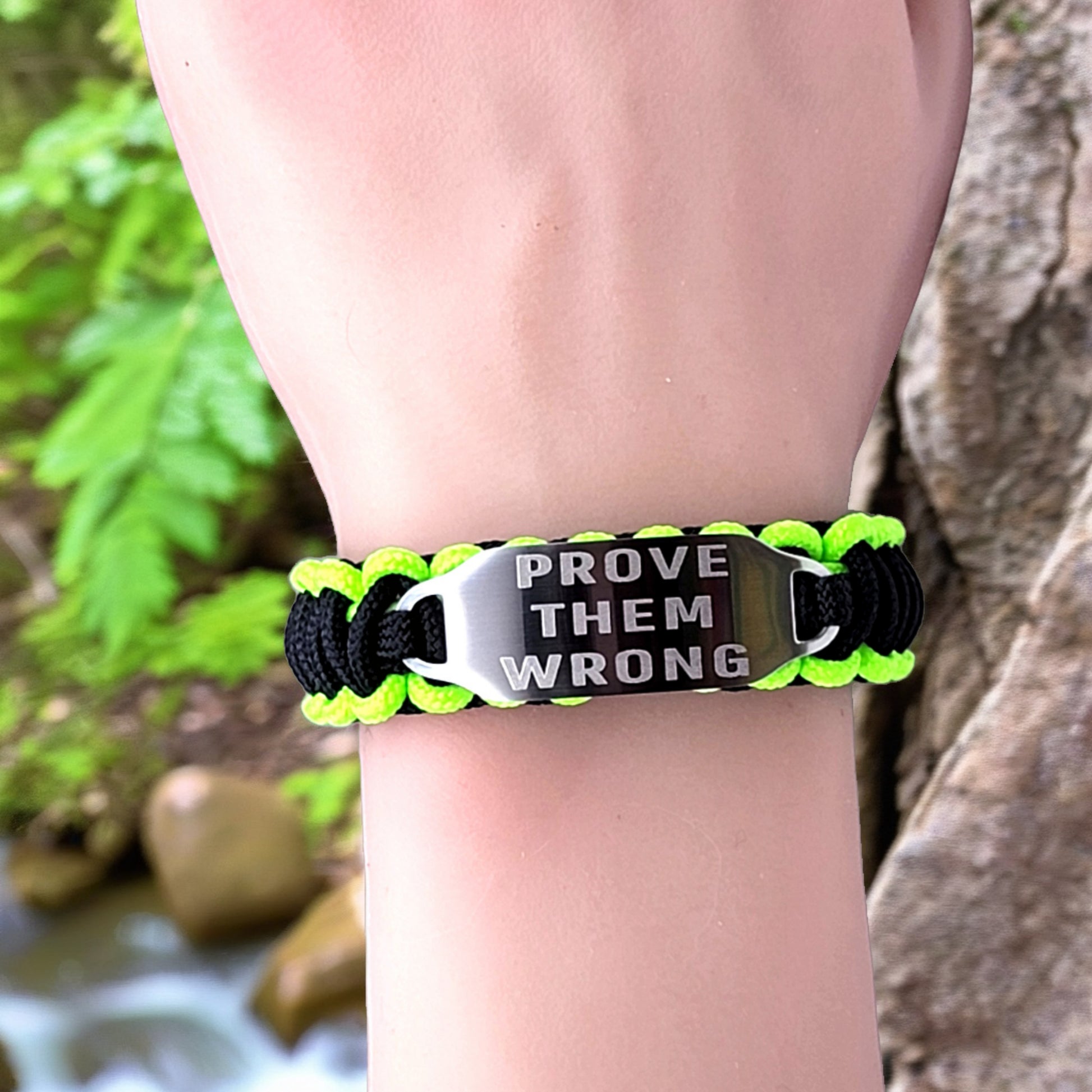 A neon green and black paracord bracelet with the phrase "Prove Them Wrong" engraved on the tag, worn on a wrist in an outdoor setting with greenery and tree bark in the background.