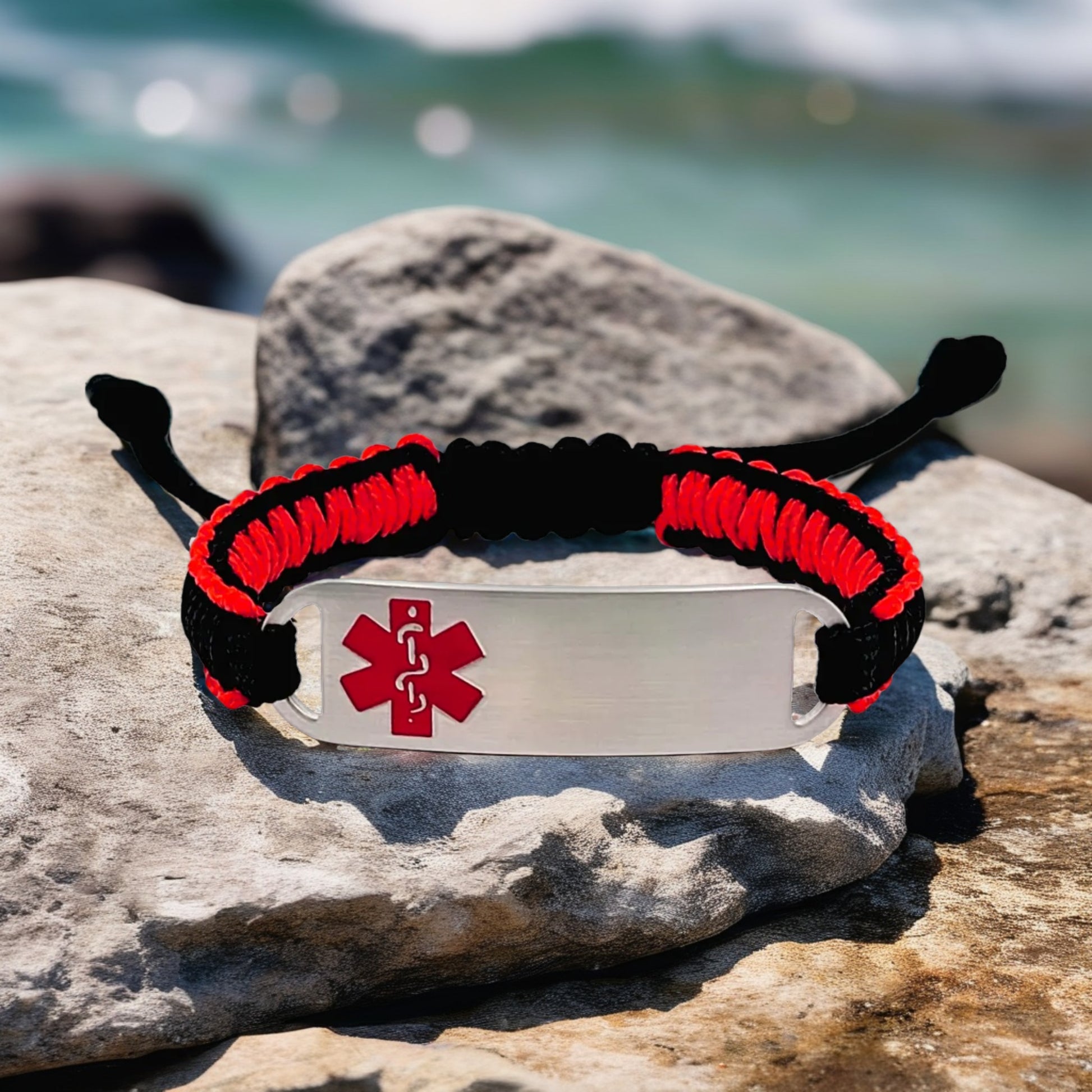 Adjustable micro paracord medical ID bracelet on a rocky surface near the ocean. Features a black and red cord design with a polished silver tag engraved with a red medical alert symbol, highlighting its bold style and functional design