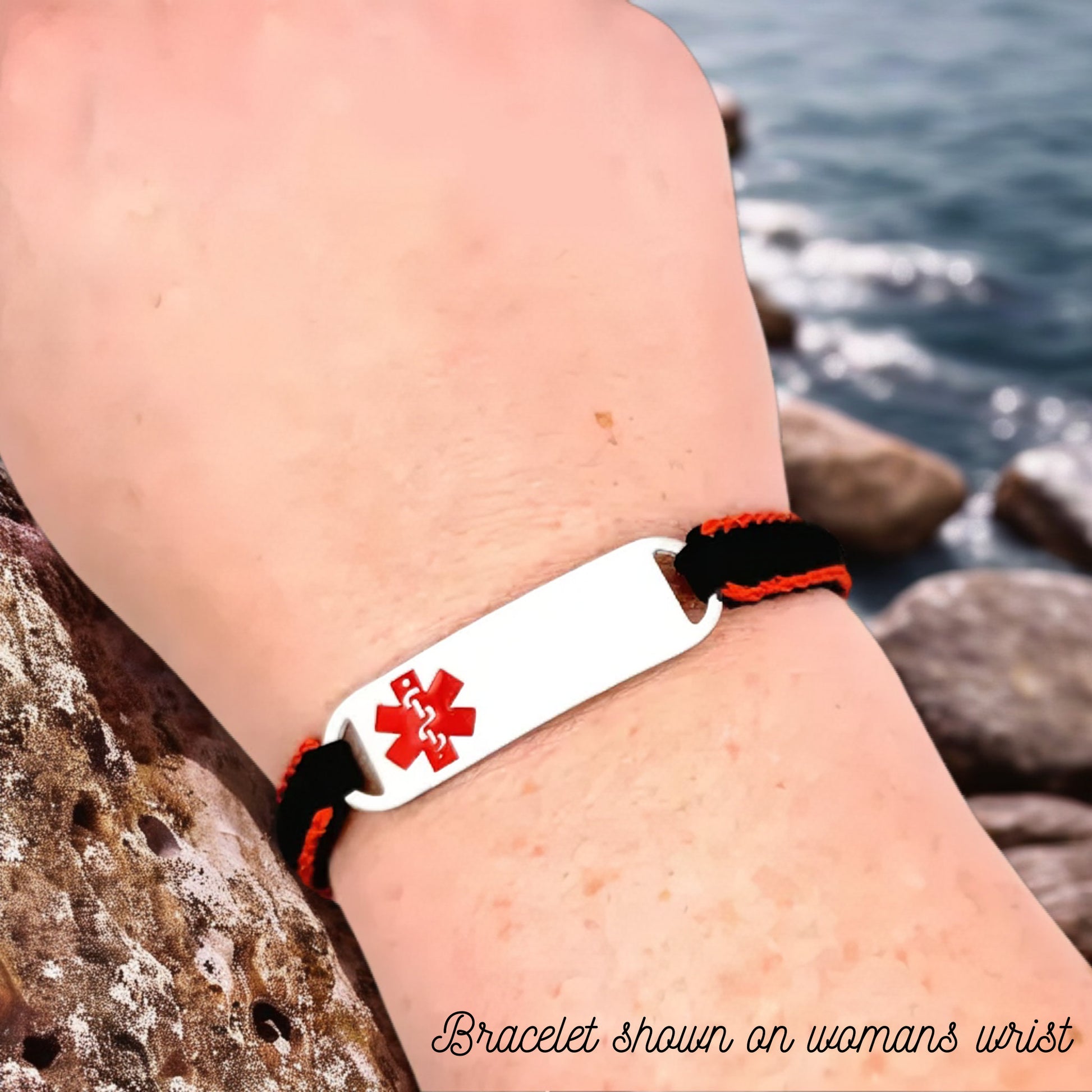 Adjustable micro paracord medical ID bracelet shown on a womans wrist near rocky water. Features a black and red cord design with a polished silver tag engraved with a red medical alert symbol, emphasizing its secure fit and bold design.
