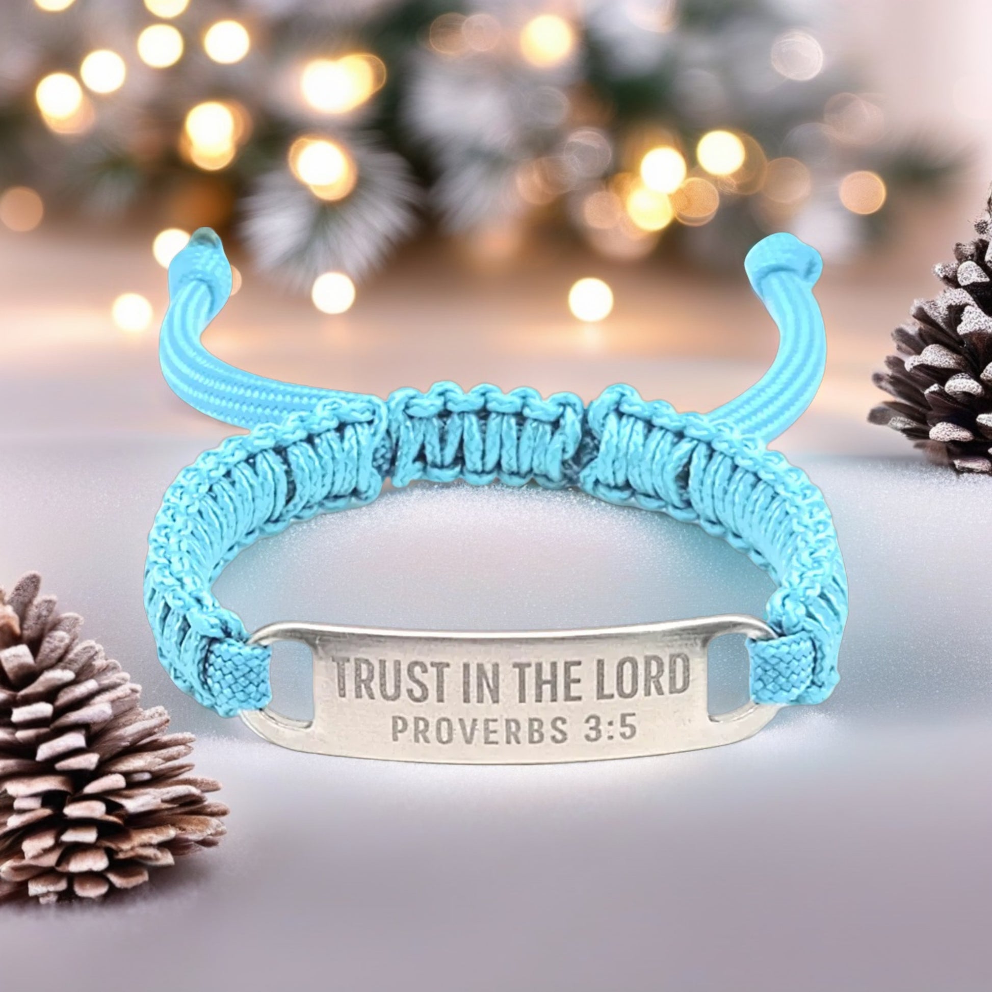 A turquoise adjustable micro paracord bracelet with a silver tag engraved with the text TRUST IN THE LORD - PROVERBS 3:5, displayed against a festive backdrop featuring softly blurred lights and pinecones