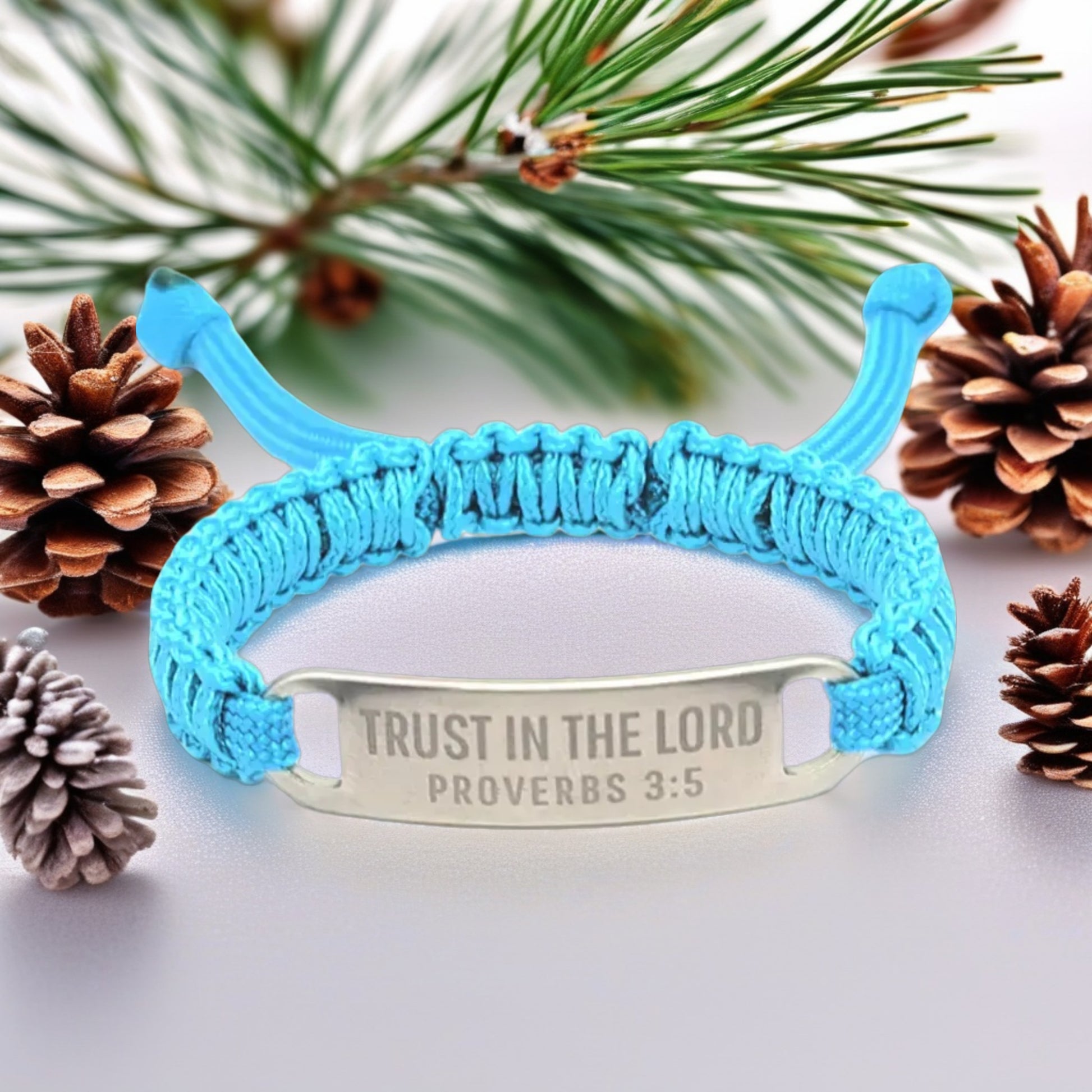 The same turquoise paracord bracelet shown against a natural backdrop of pine needles and pinecones, highlighting the engraved tag with the scripture verse.