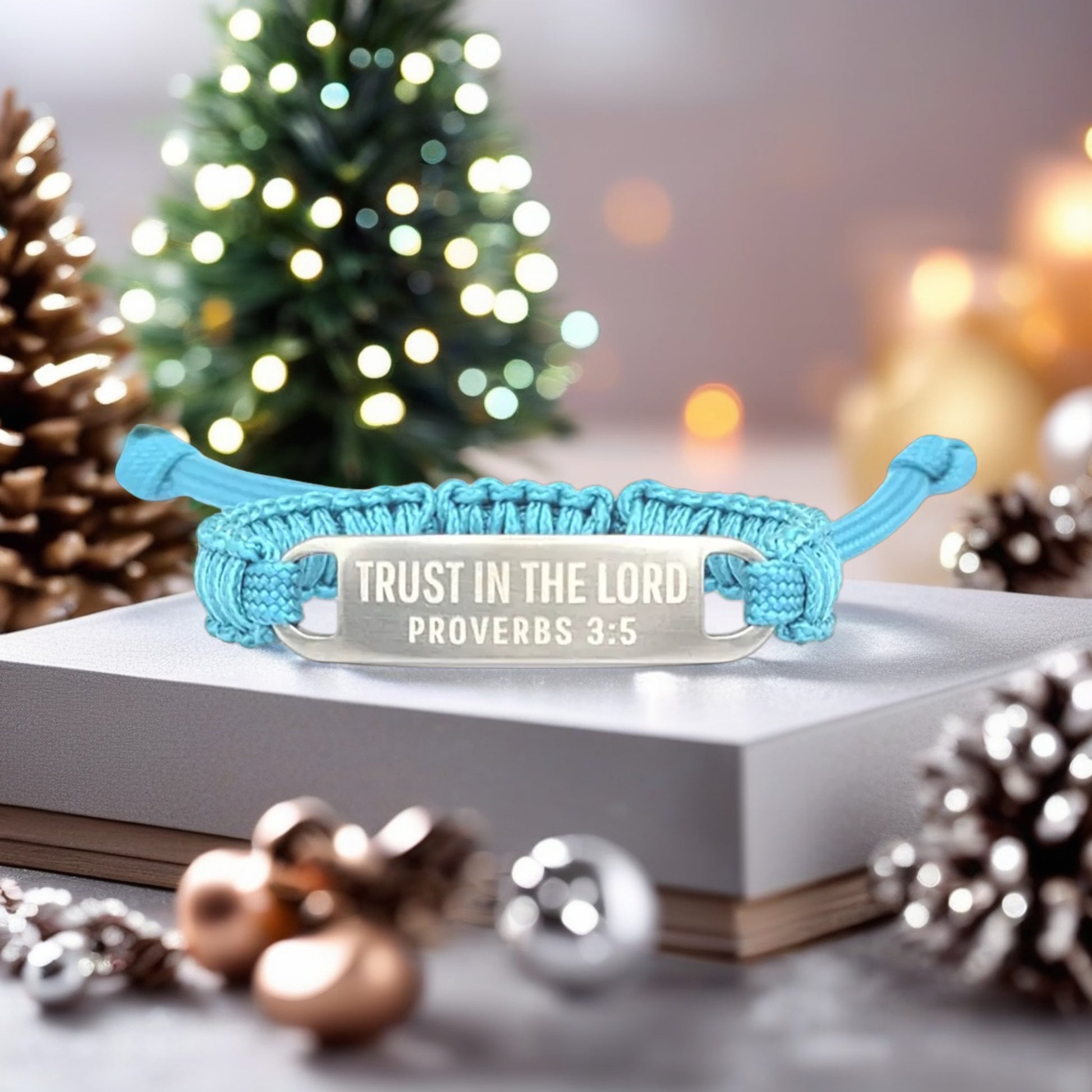 A close-up of the turquoise scripture bracelet resting on a white surface, surrounded by Christmas decorations, including ornaments and a lit tree in the background.