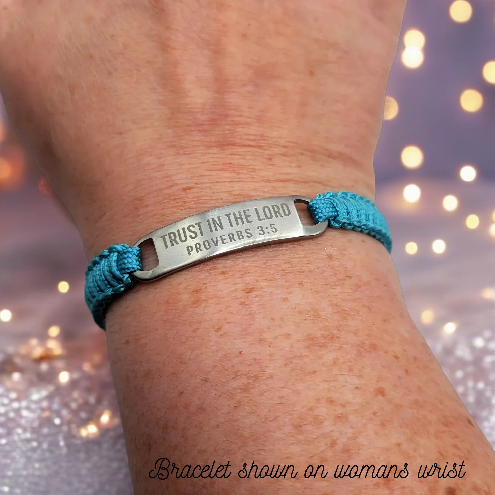 The turquoise bracelet worn on a woman's wrist, showing the engraved TRUST IN THE LORD - PROVERBS 3:5 tag up close with a festive blurred background of soft golden lights