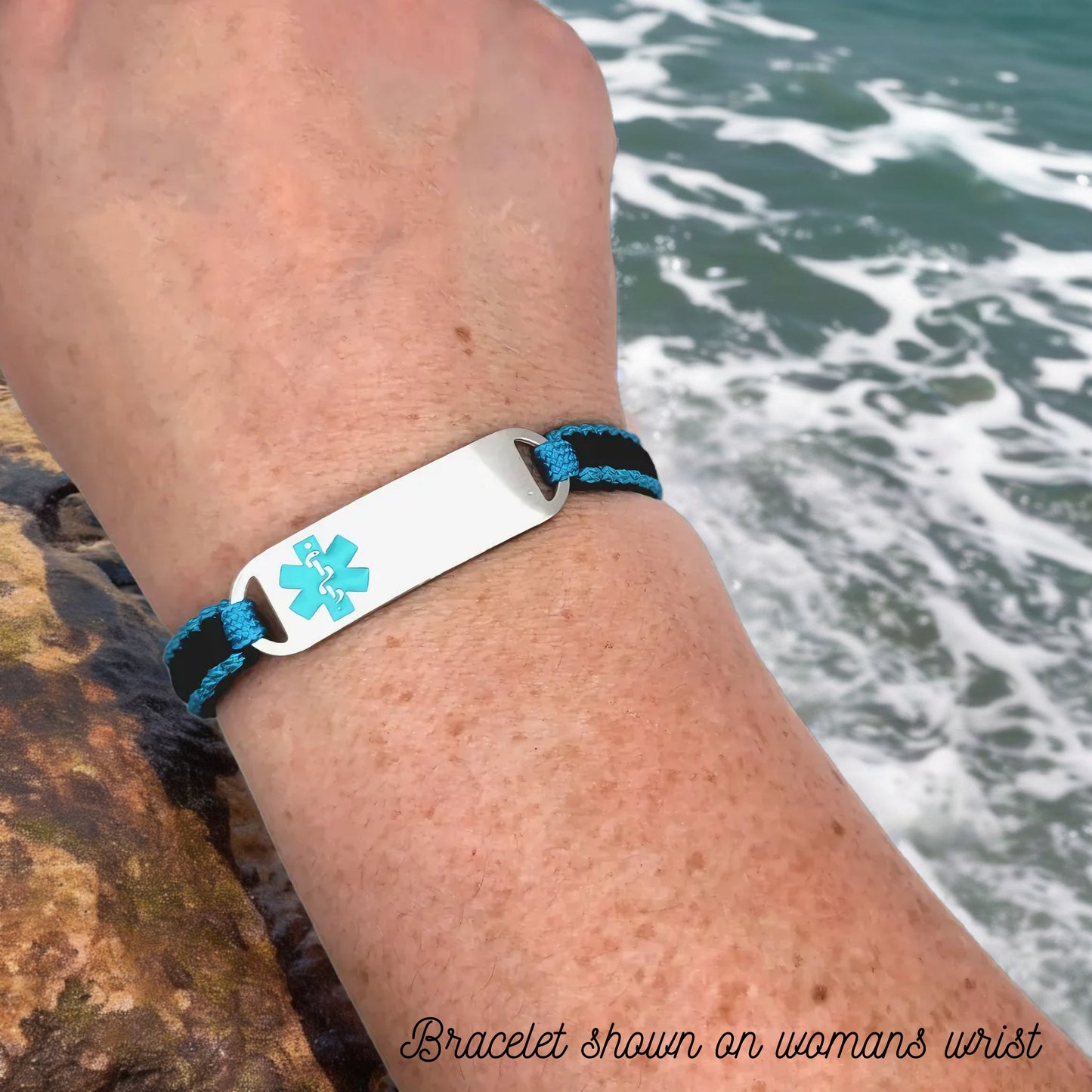 Adjustable micro paracord medical ID bracelet shown on a womans wrist near ocean waves. Features a navy and teal cord design with a polished silver tag engraved with a teal medical alert symbol, emphasizing its secure fit and stylish appearance.