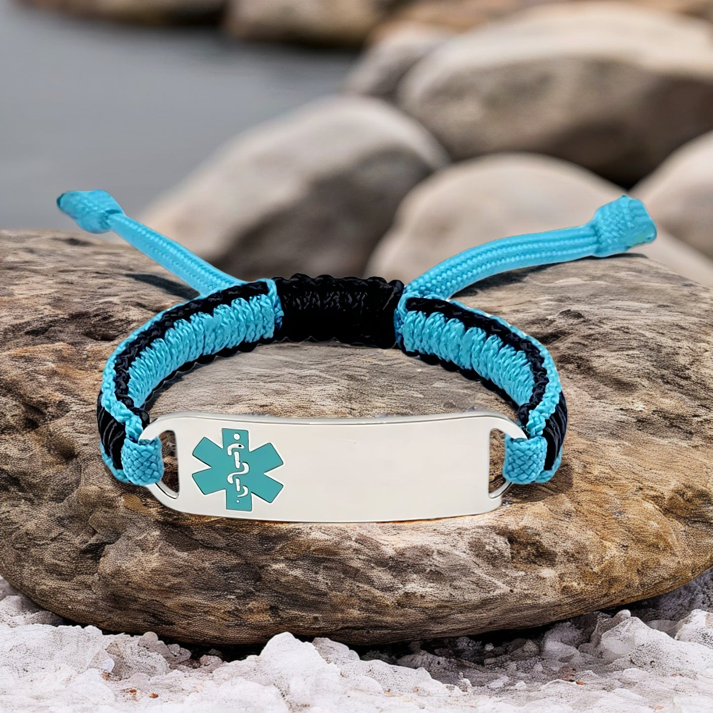Adjustable micro paracord medical ID bracelet on a rocky surface near water. Features a navy and teal cord design with a polished silver tag engraved with a teal medical alert symbol, highlighting its stylish and functional design.