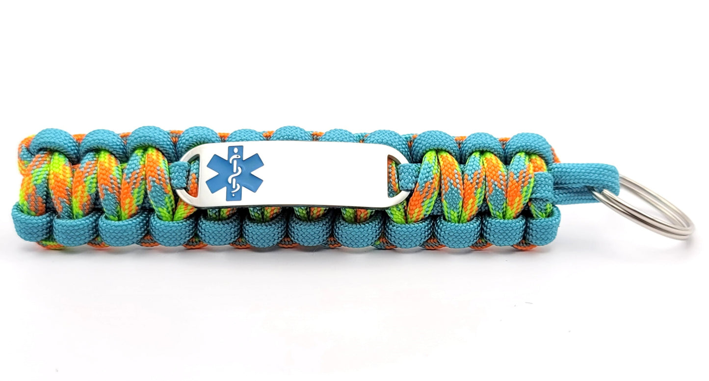 Brightly colored light blue and multicolored paracord medical ID keychain with a silver, stainless steel ID tag with an ocean blue enameled medical symbol, displayed on a white background.